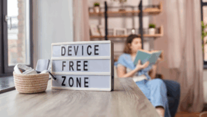 Sign saying Device Free Zone women blurrewd in the background reading a bvook her devises in a basket next to the sign