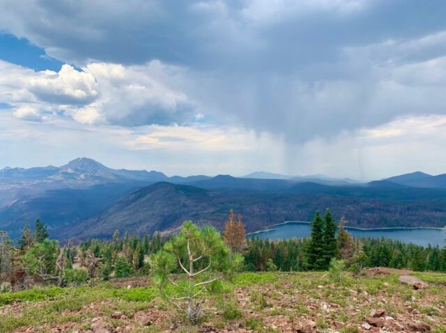 A mountainous landscape with a lake, trees, and a cloud-filled sky.